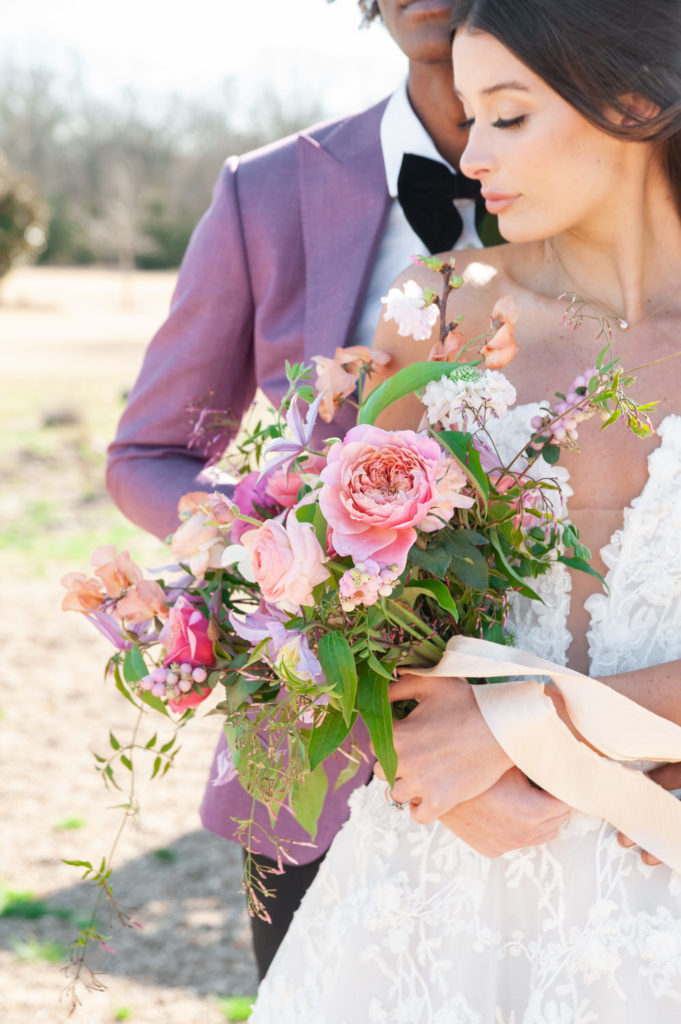 bride holding a pink and purple floral bouquet with her groom behind her in the countryside of Quinlan Texas