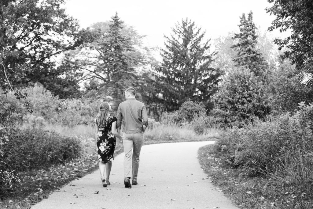 man and woman holding hands and walking down a paved trail during their summer engagement photos in grand rapids