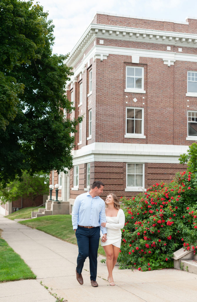 couple walking and holding hands in front of buildings and a red rose bush