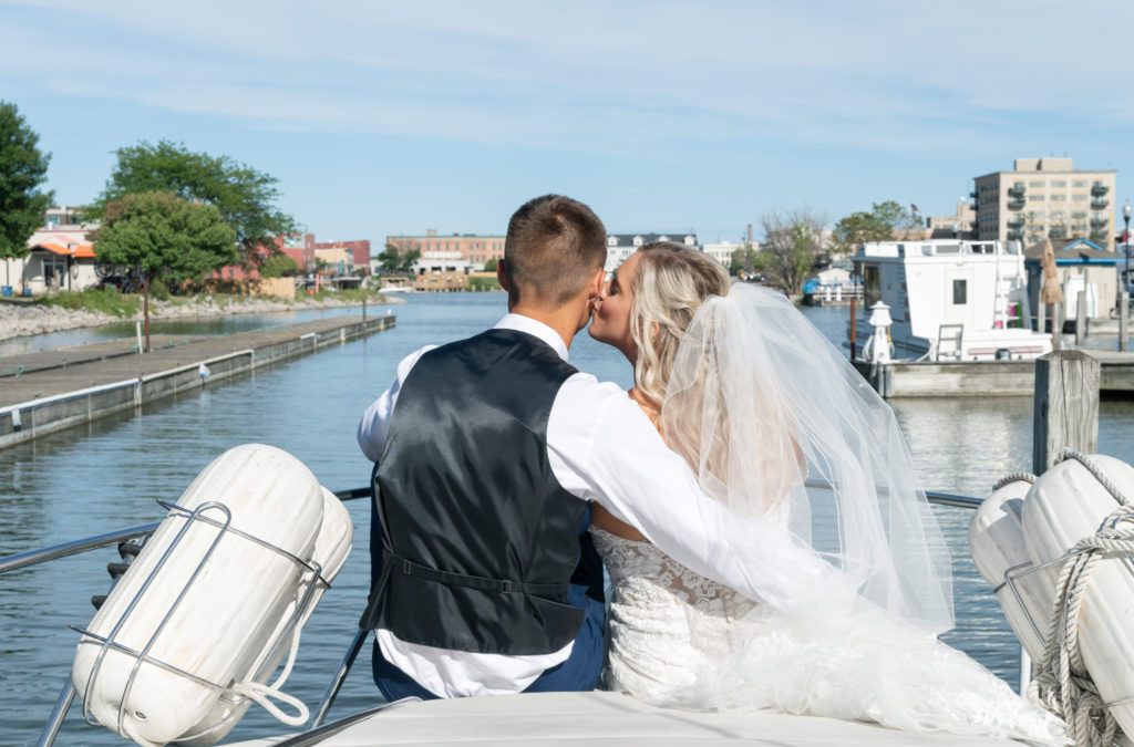 bride kisses the groom's cheek while they both sit on a yacht with the river in front of them