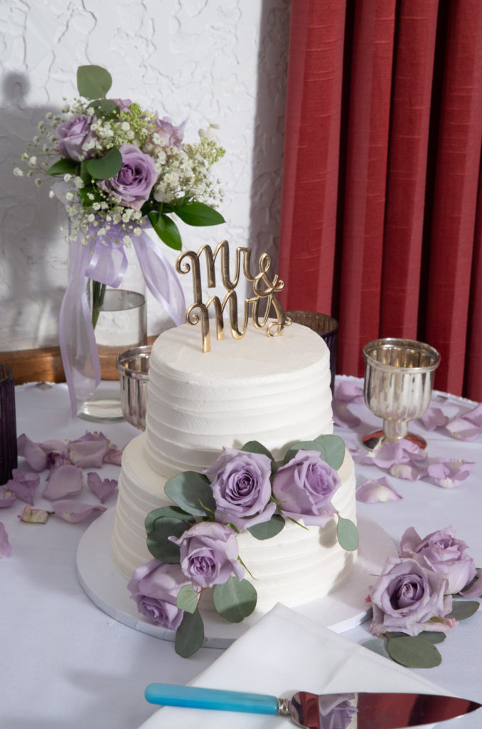 white two layer wedding cake with purple flowers draping it on the side
