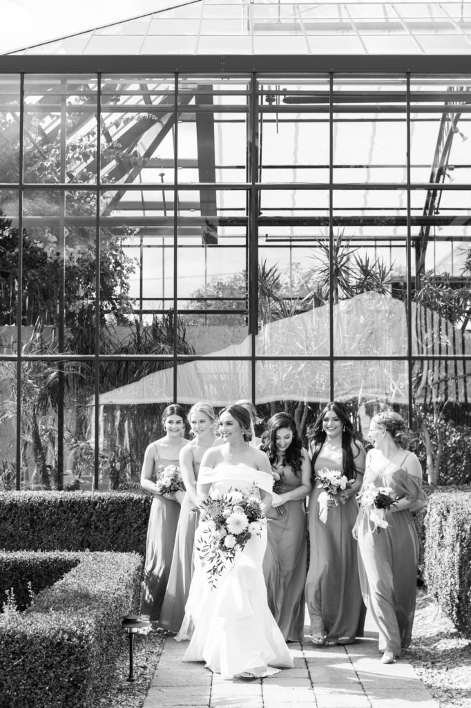 bridesmaids walk behind the bride in front of a modern greenhouse