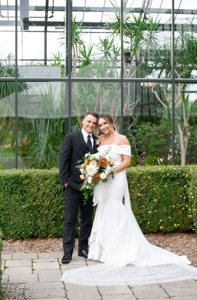 Dalton and Alyssa smile on their wedding day with a modern greenhouse behind them