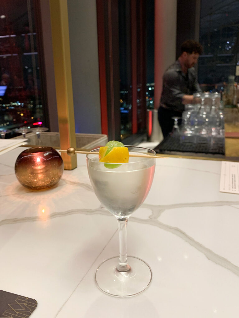 a crisp and clear cocktail sitting on a marble bar top at night with city lights in the background