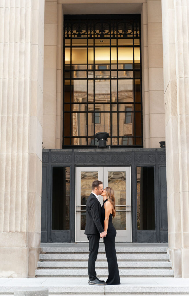 man and woman in black formal clothing kiss in front of the doors of the Civic Auditorium 