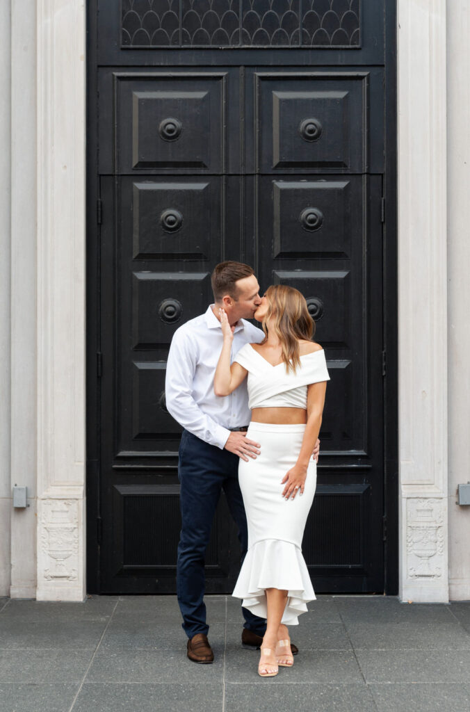 man and woman sharing a kiss in front of the black arch door of the Amway Grand Plaza during their Grand Rapids engagement photo session