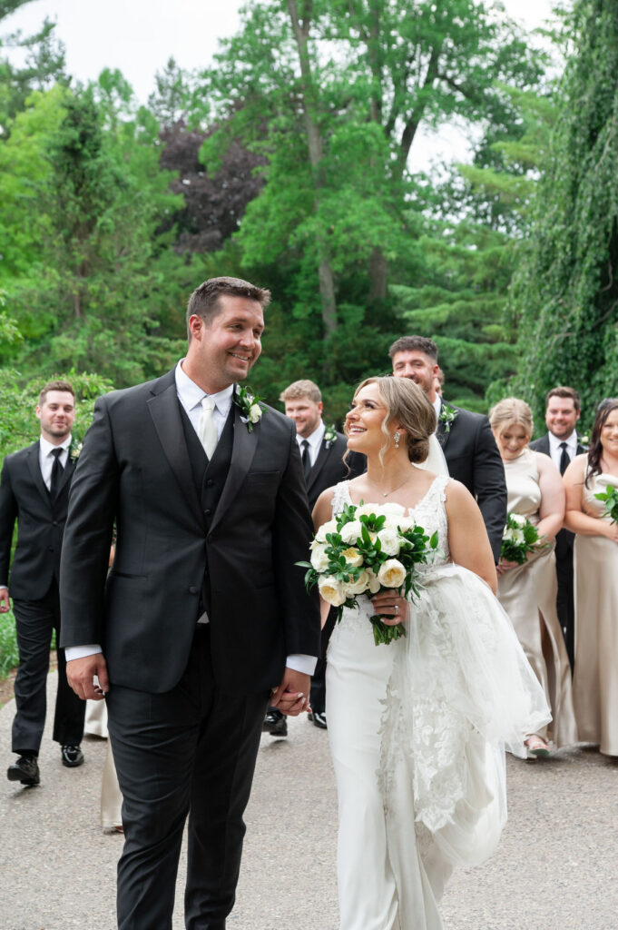 Bride and groom walking hand in hand with the wedding party walking behind them during photos at Dow Gardens in Midland, Michigan
