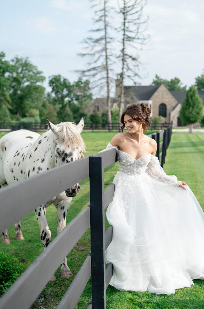 bride petting a white horse with black spots on her summer wedding day at Venue3Two