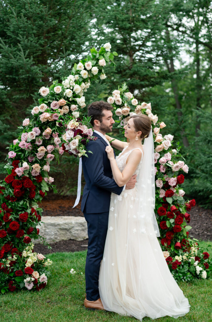 bride and groom embracing each other in front of a floral wedding ceremony arbor made up of red, pink and white garden roses