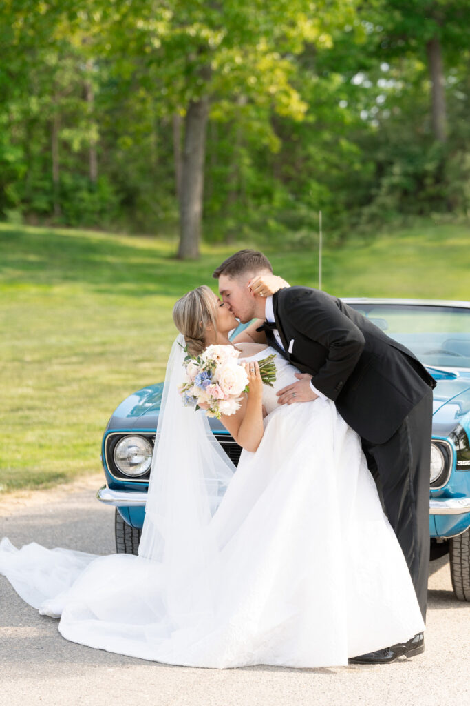 groom dips and kisses his bride in front of a blue camaro at their Waldenwoods wedding in the spring