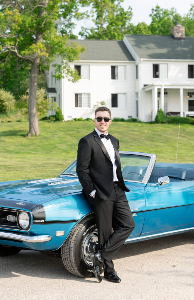 groom leaning up against a vintage blue Chevrolet Camaro smiling with black sunglasses
