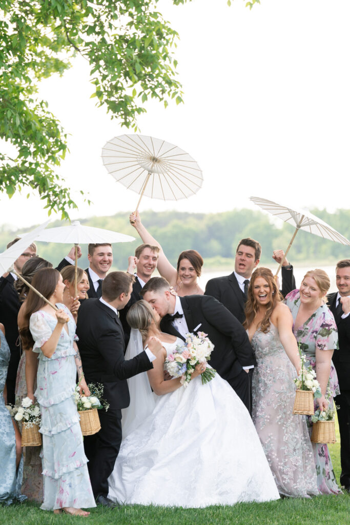 bride and groom kiss as their bridal party surrounds them and cheers in front of the lake at Waldenwoods