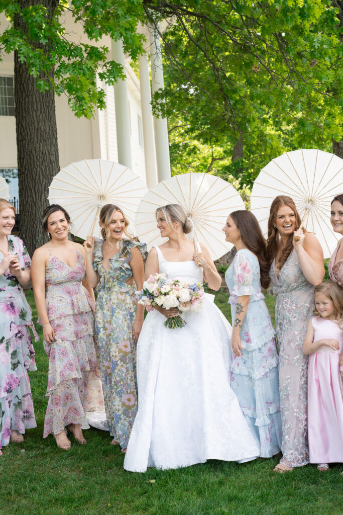 bride smiling at all her bridesmaids in their pastel various pattern dresses as they all old white paper parasols at Waldenwoods in Howell, MI