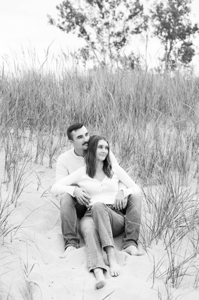 engaged couple sitting together in the sand dunes looking out onto Lake Michigan in Muskegon