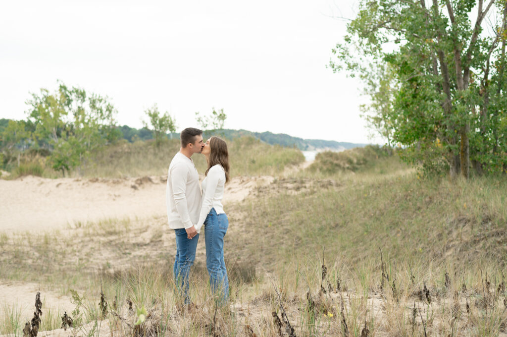 Becca and Alex holding hands and kissing with Lake Michigan sand dunes and water in the background