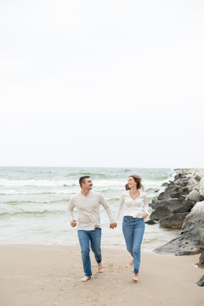 man and woman holding hands and running together during their Muskegon engagement session on Lake Michigan
