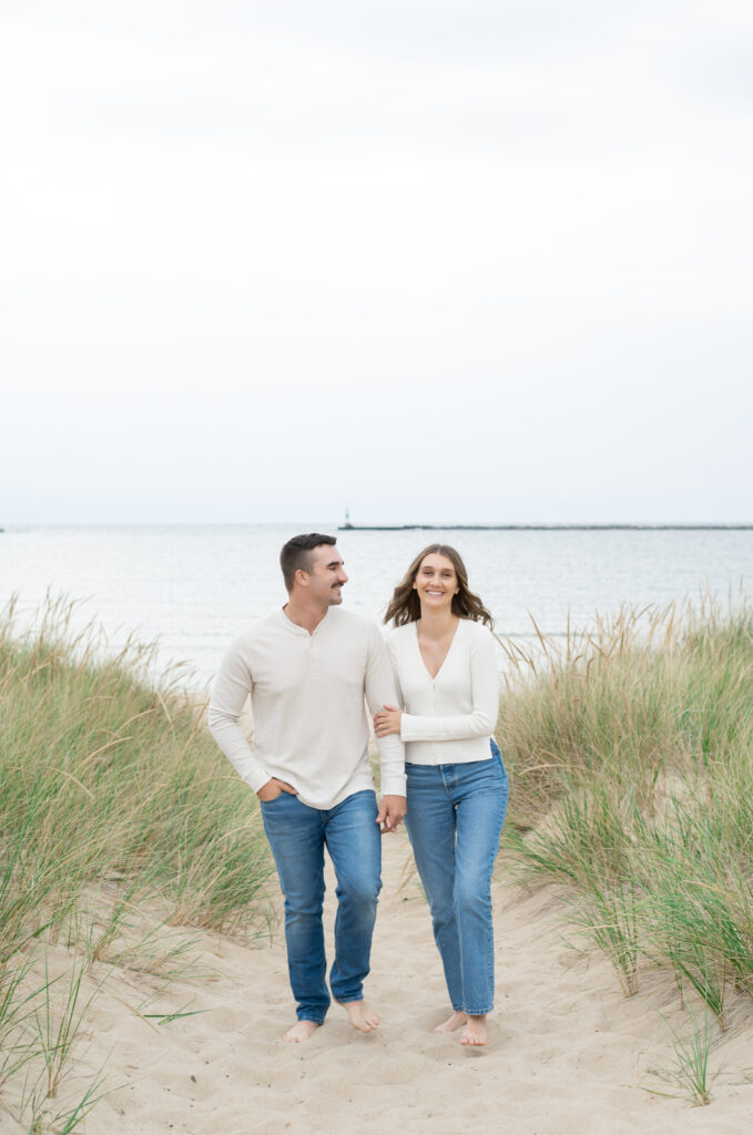 engaged man and woman holding hands and walking on the sand dunes with the Lake Michigan shoreline in the background