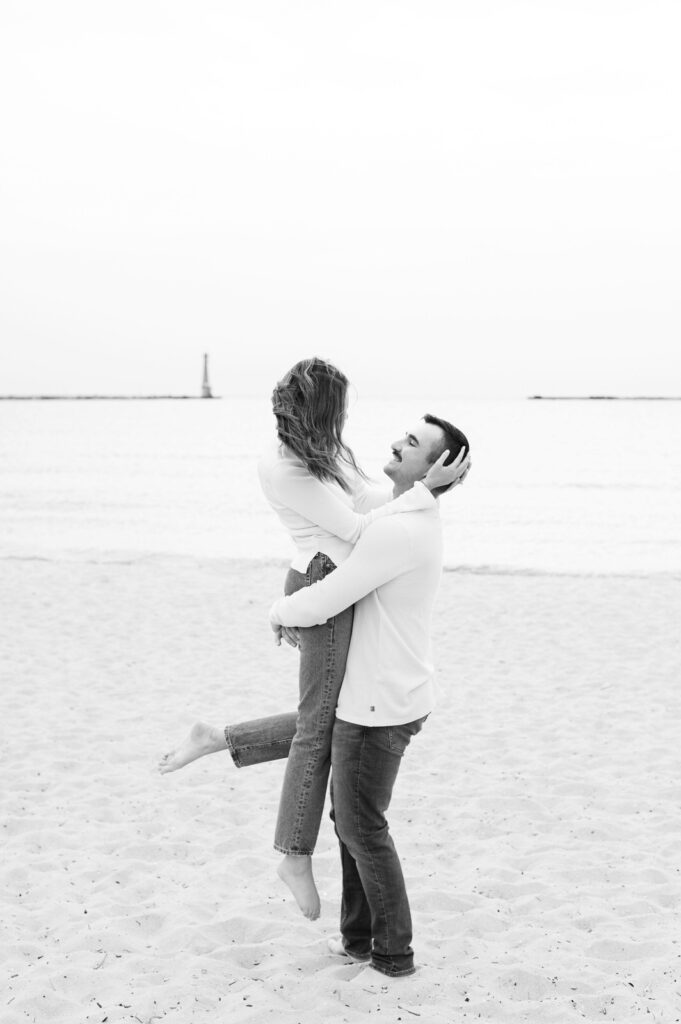 man holding up his fiance in his arms on the shore of Lake Michigan