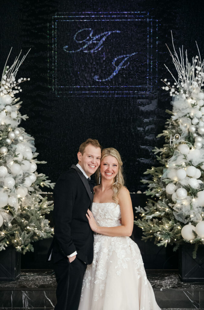 bride and groom posing and smiling in front of their custom monogram waterfall and Christmas trees