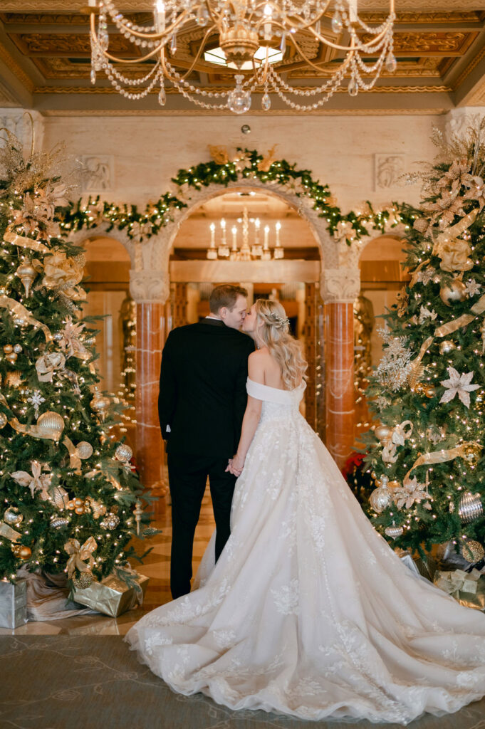 bride and groom kiss in the entrance of Saint John's Resort in Plymouth, MI with gold decorated Christmas trees on each side of them