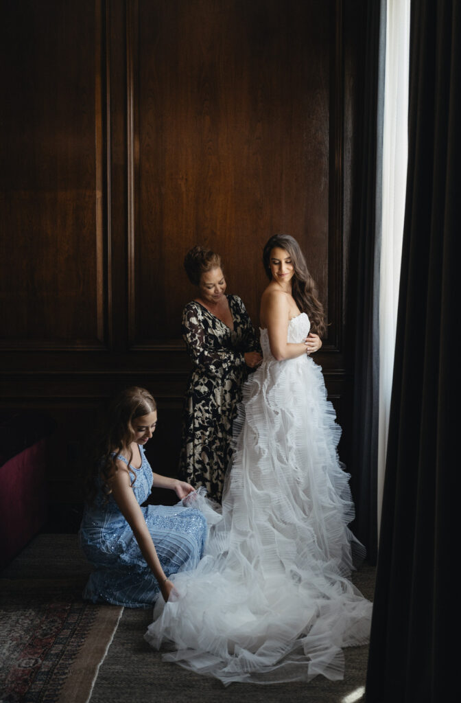 mother of the bride zips up her wedding dress while her sister adjusts the end of her dress in front of a window in a suite at the Detroit Foundation Hotel