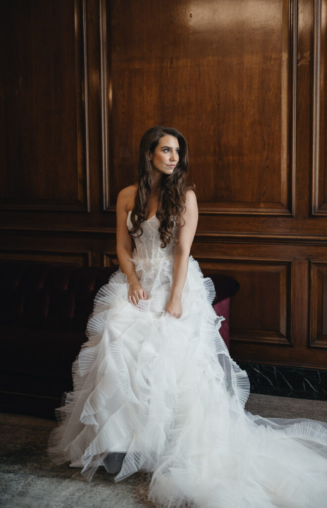 bride sitting on the arm of a burgundy velvet couch while looking out the window of her suite in the Detroit Foundation Hotel