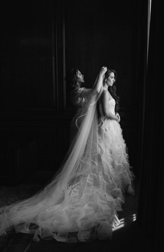 bridesmaid places a long veil on the bride's head as she looks out the window of her suite at the Detroit Foundation Hotel