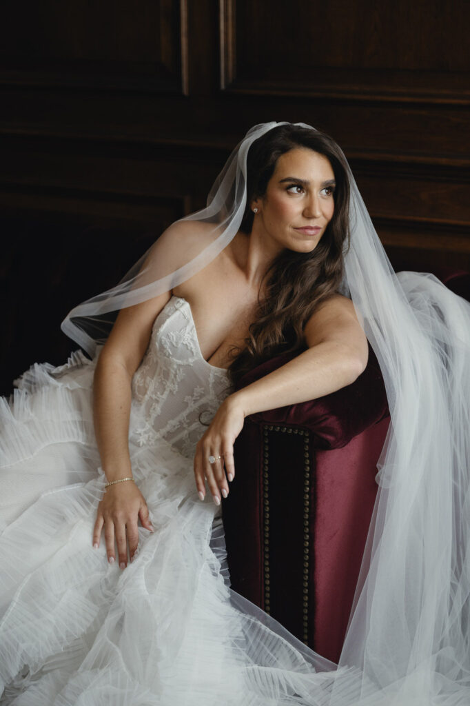 bride sitting in a burgundy velvet couch while leaning against the arm rest and gazing out a window of the Detroit Foundation Hotel