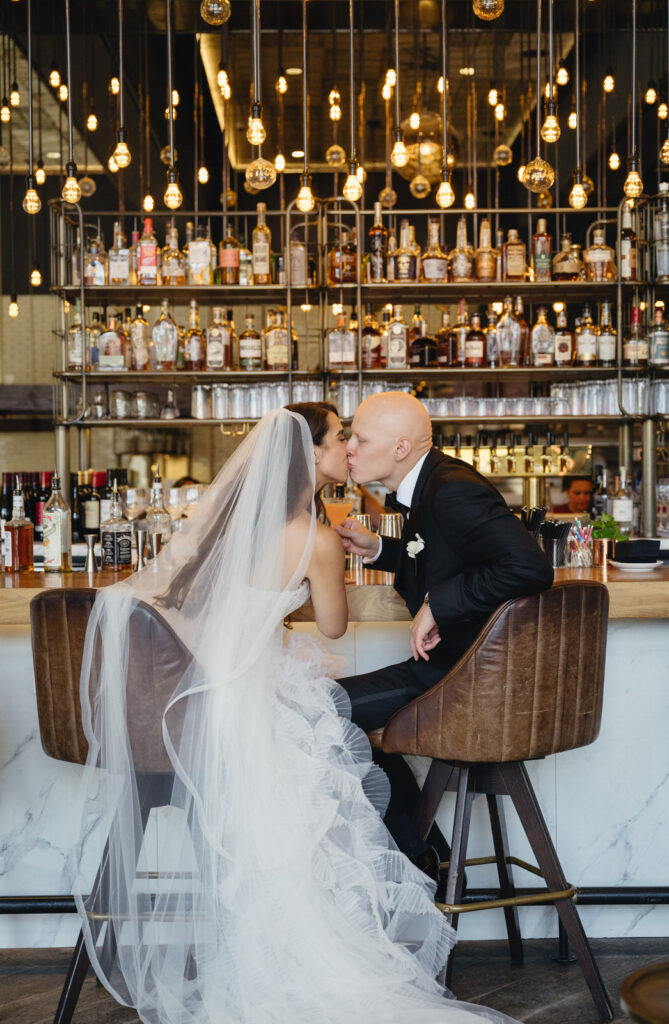 bride and groom sitting in the brown leather barstools while kissing at the bar in the Detroit Foundation Hotel