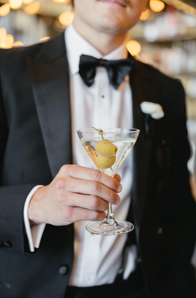 man in a black suit and black bowtie holds a martini with 2 olives at the bar inside the Detroit Foundation Hotel