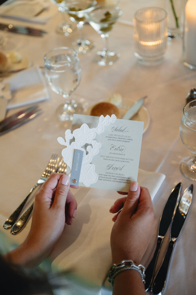 guest holds a custom menu design by Alyssa Amez featuring dusty blue, white, and gray colors with an embroidered floral pattern