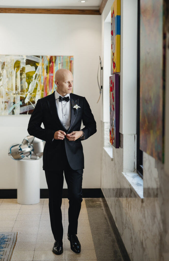 groom buttoning up his black suit coat while walking by a window in the Detroit Foundation Hotel