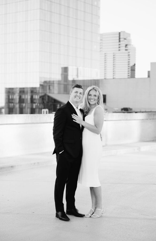 man in a black suite and his fiance standing and smiling with the Grand Rapids city skyline behind them at their West Michigan engagement session