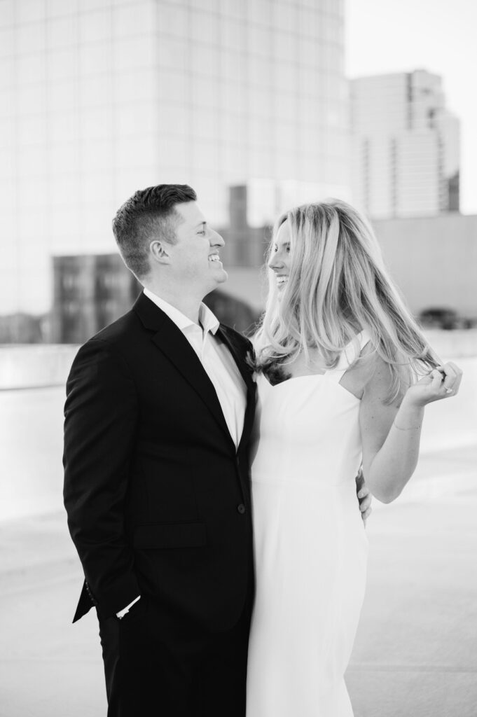 couple smiling at each other with the Grand Rapids city skyline behind them at their West Michigan engagement session