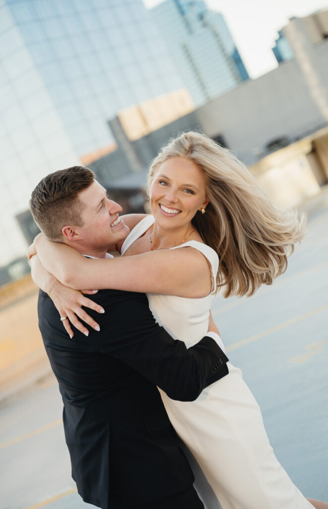 engaged man holding his fiance around her waist as she smiles with her hair blowing in the wind on top of the Amway Grand Plaza parking ramp