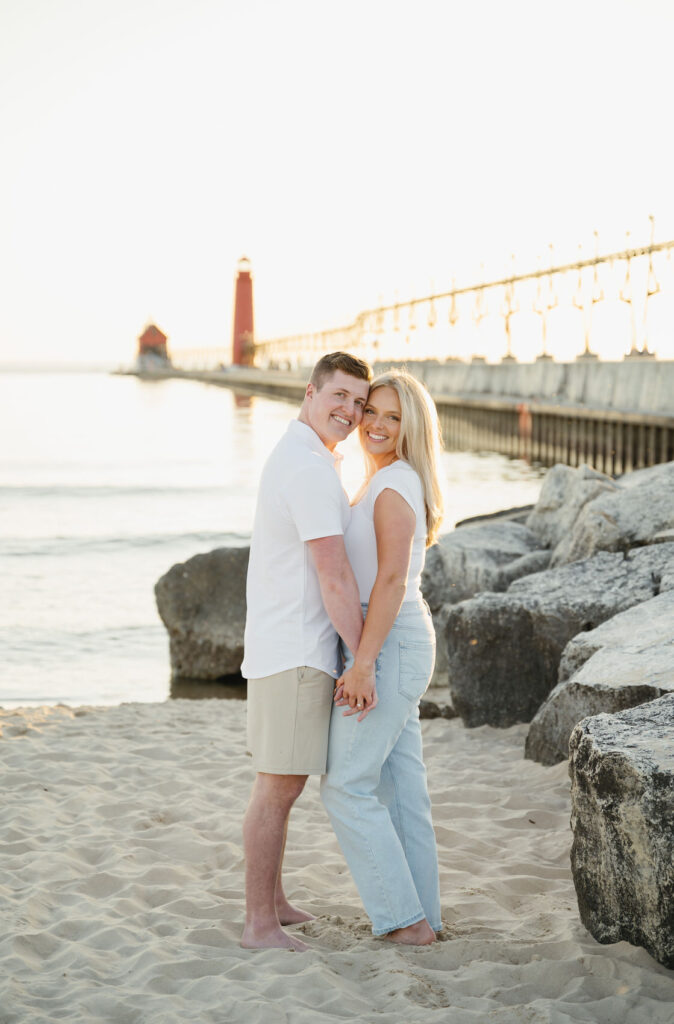 engaged couple holding hands and chest to chest in front of the Grand Haven State Park Pier at sunset