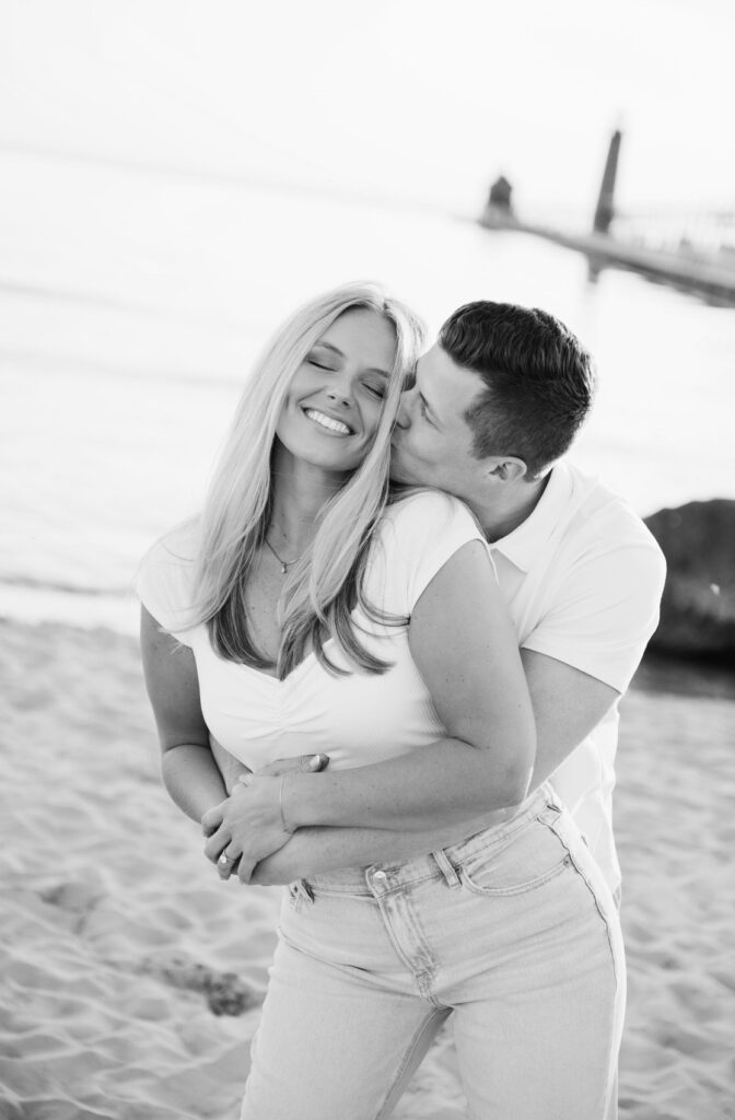 man kisses the cheek of his fiance while hugging her from behind during their west Michigan engagement session
