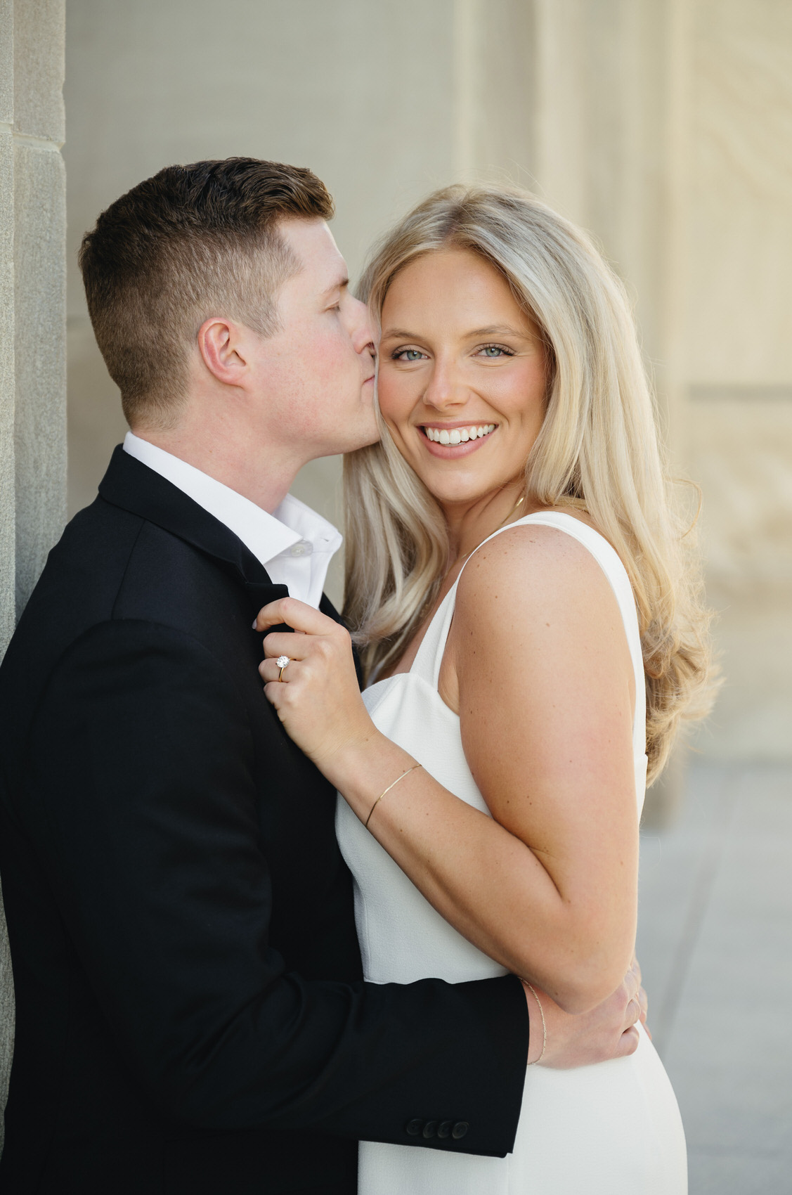 man in a black suit giving his fiance in a white dress a kiss on her cheek