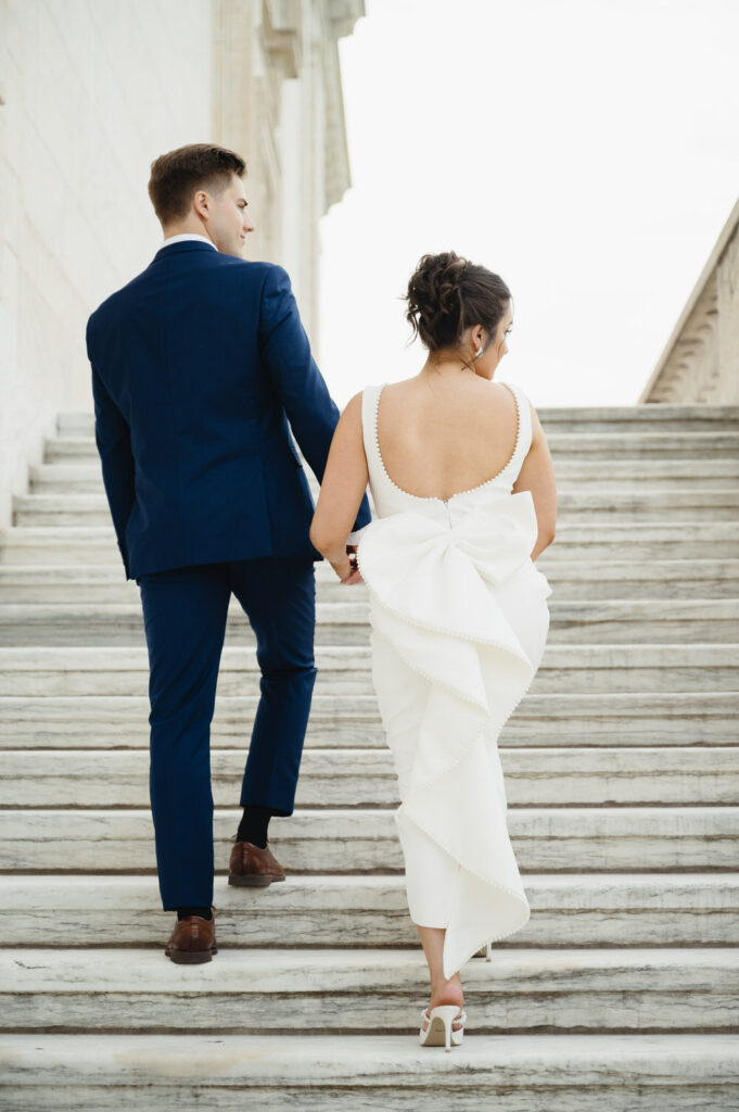 engaged couple in a white dress and blue suit walk up the marble stairs of the Detroit Institute of Arts
