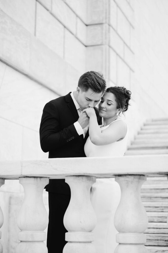 engaged man kisses his fiance's hand as she smiles down on the steps of the Detroit Institute of Arts